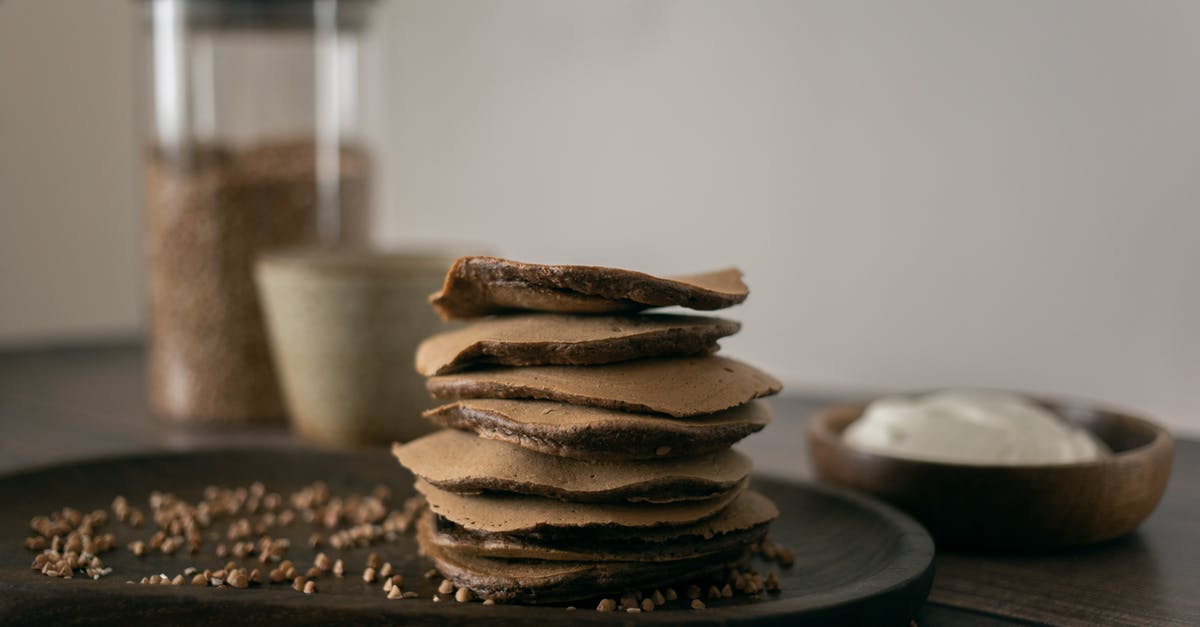 Sauce bubbling out of jar - Healthy buckwheat pancakes on plate with scattered grains placed on wooden table with sour cream in light kitchen at home