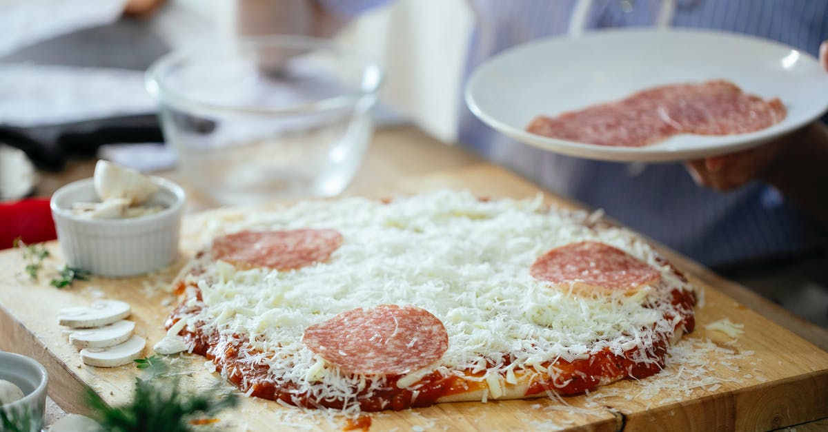 Separate cutting boards: Cooked vs Uncooked meat - Crop anonymous person standing near table with uncooked homemade pizza and adding slices of salami while preparing in kitchen at daytime