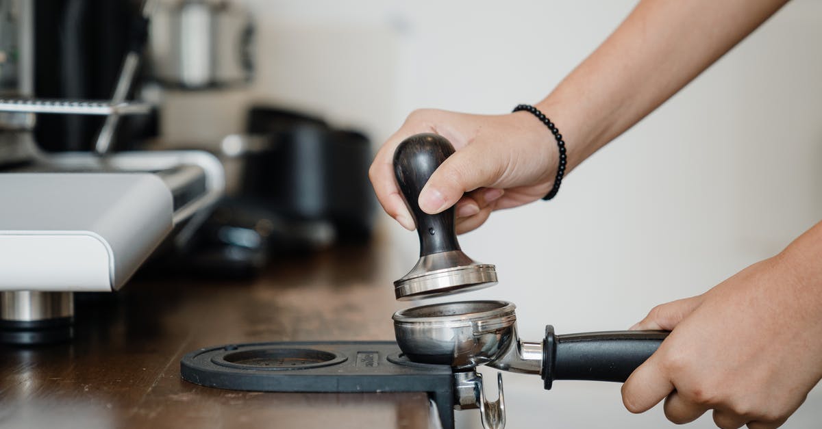 Steam "nature" of cappuccino steamers - Crop anonymous barista pressing aromatic coffee beans into filter block by using tamper while standing in light kitchen nearby professional brewing machine