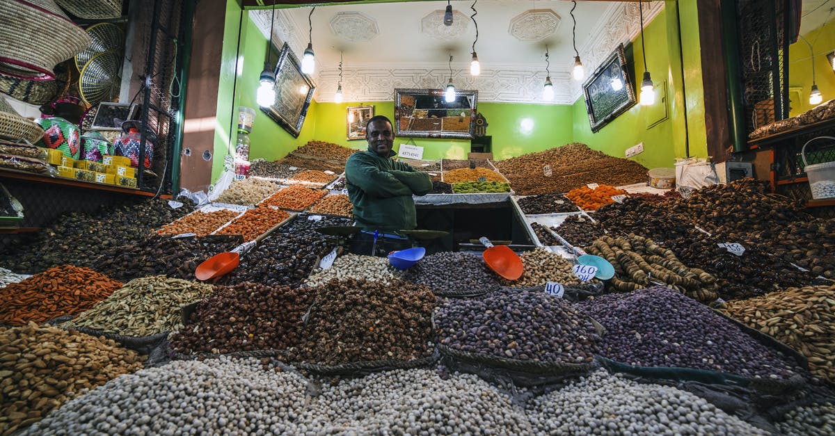 Storing/transporting nut brittle in tins - From below of African American man standing between market stalls with multi colored dried fruits and nuts illuminate with light bulbs