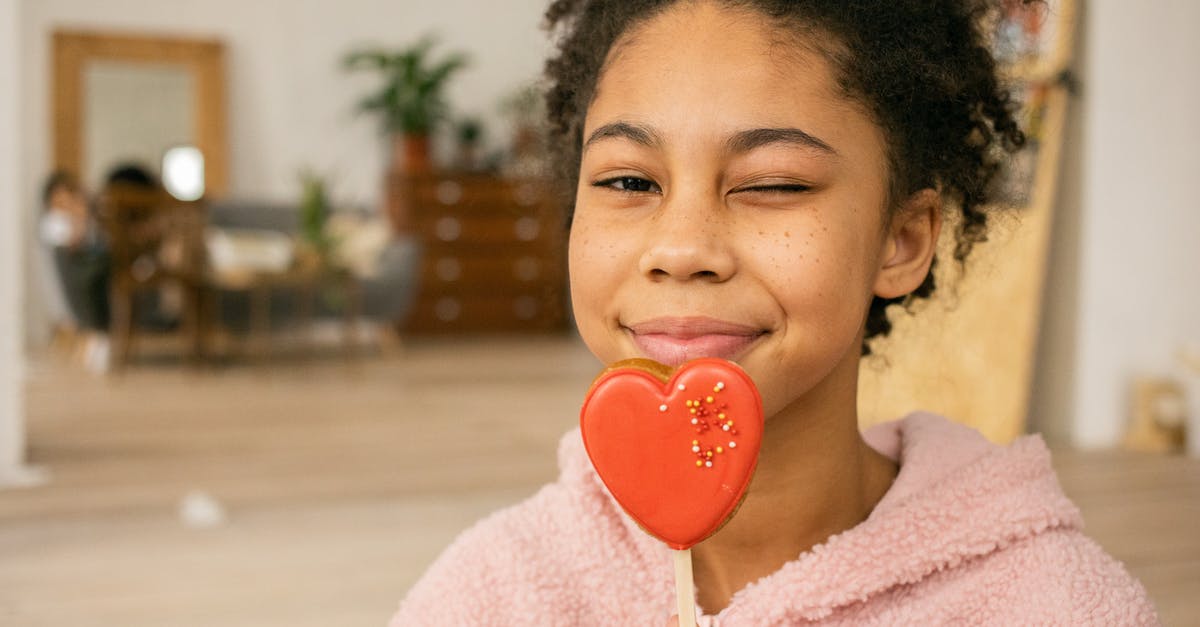 Sugar content of food - Happy African American teenage girl with sweet heart shaped gingerbread cookie on stick winking and looking at camera in light room on blurred background