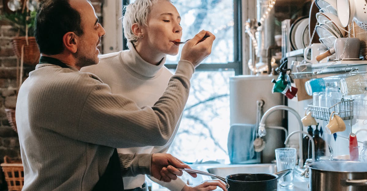 Tasting food in kitchen - Side view of content ethnic man in casual clothes and apron feeding happy wife while preparing delicious dinner at stove in kitchen