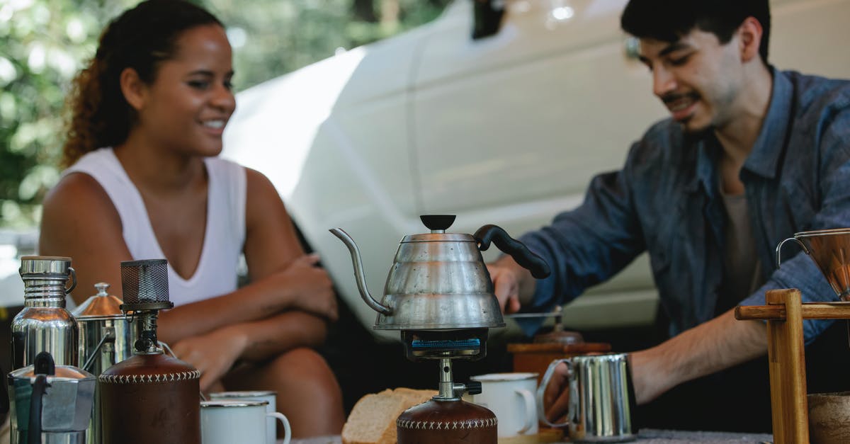 Time of cooking: induction stove vs gas stove - Young cheerful couple in casual wear boiling water in kettle on gas stove and grinding beans for coffee brewing while spending sunny day in camp