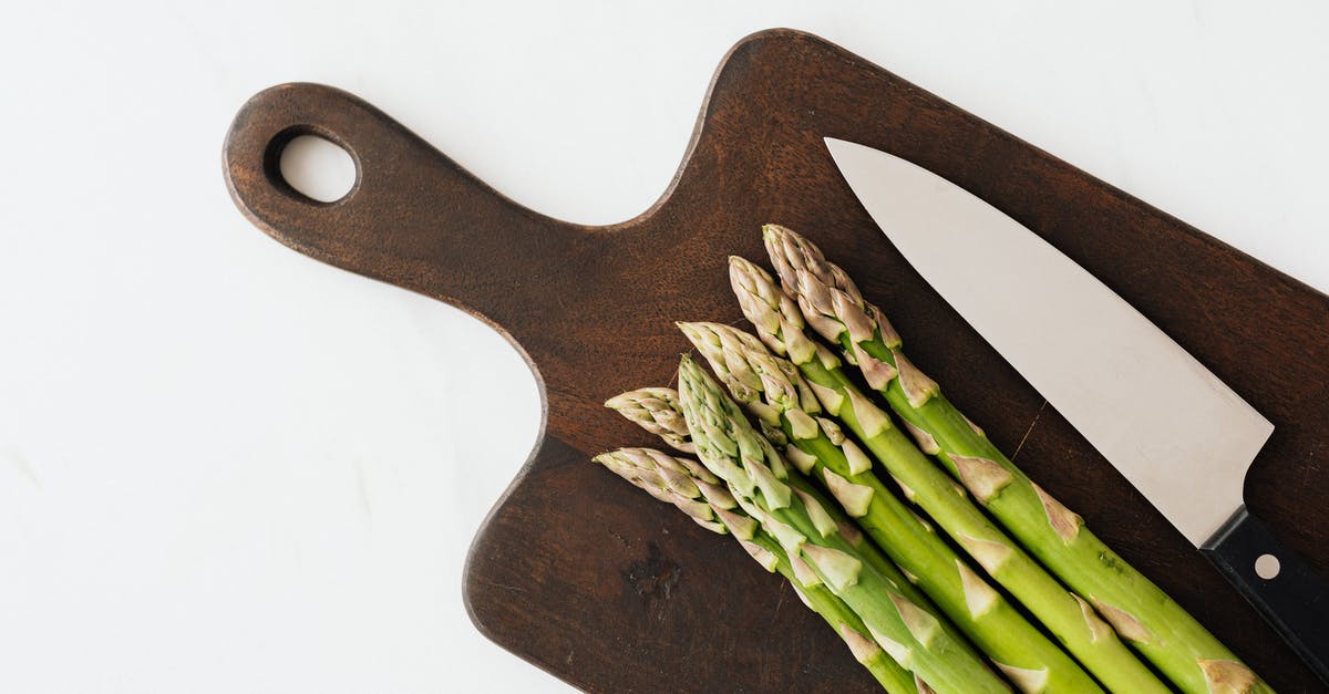 Transfer vegetables objects from cutting board to pan - Top view of bunch fresh green asparagus with knife on dark brown cutting board placed on white surface