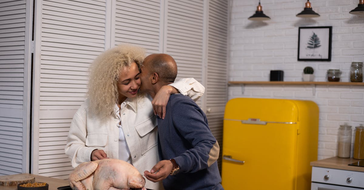 Turkey stock in refrigerator - Tender African American husband kissing positive black wife with closed eyes while standing near counter with uncooked turkey during cooking in modern kitchen