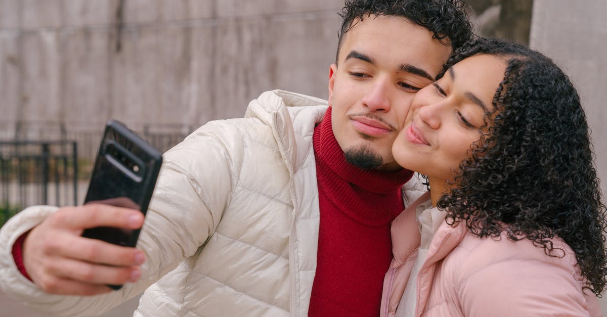 Use by date on cacao butter - Enamored young Hispanic couple in warm casual clothes smiling and touching cheeks while taking selfie on mobile phone standing on street