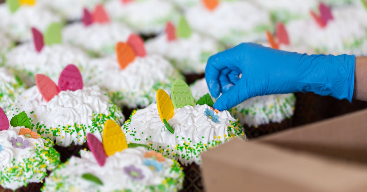 Using manual egg beater to cream eggs and sugar - Crop unrecognizable worker in disposable glove with box against tasty desserts with egg and floral decor on whipped meringue cream