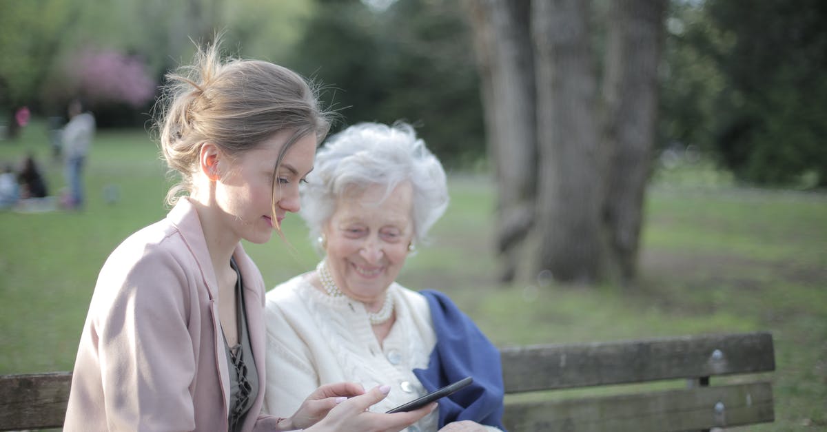 Using old vanilla beans - Side view of smiling adult female helping aged mom in using of mobile phone while sitting together in park