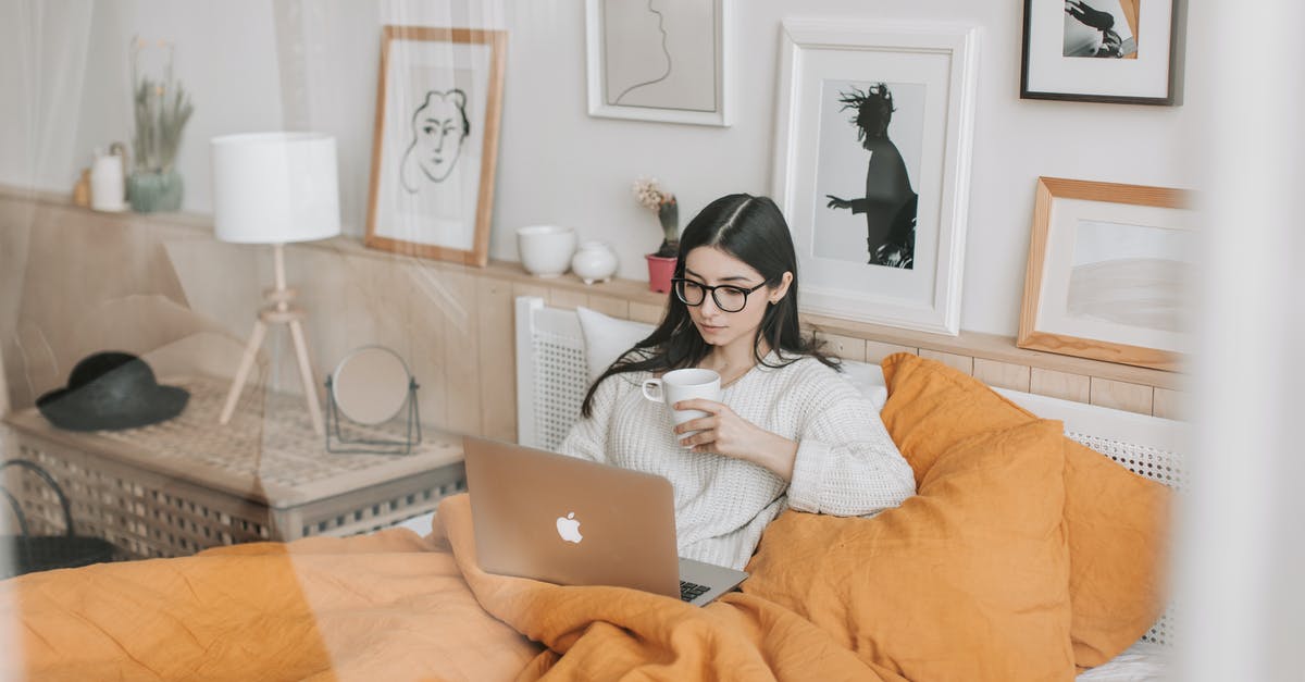 Using sous vide to warm canned vegetables - Focused young brunette in eyeglasses with cup of hot drink lying in comfortable bed and working on laptop in morning