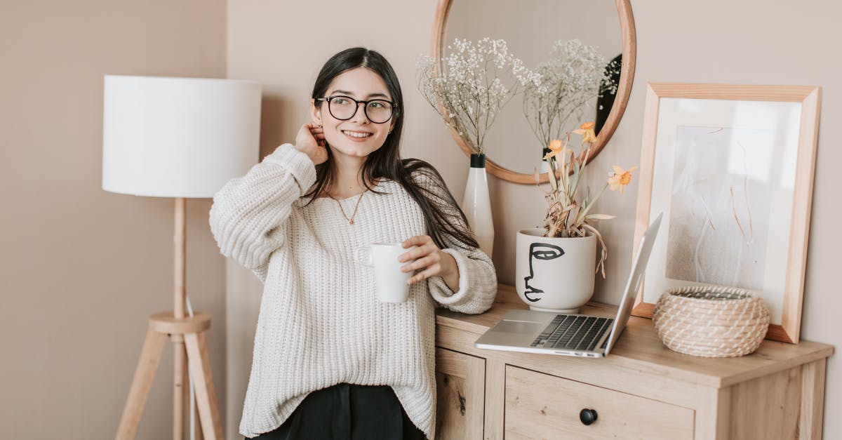 Using sous vide to warm canned vegetables - Dreamy smiling young lady in white sweater and eyeglasses with mug of hot beverage looking away happily while standing near bedroom dresser and browsing laptop