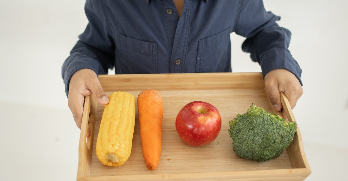What's the difference between broccoli from China and broccoli from Australia? - From above of crop anonymous kid in casual outfit holding wooden tray with fresh healthy broccoli apple carrot and corn against white background