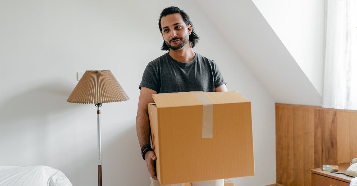 What's this odd stuff coming out of my meringue? - Cheerful young bearded male in casual wear carrying cardboard container in sunny bedroom while moving into new apartment