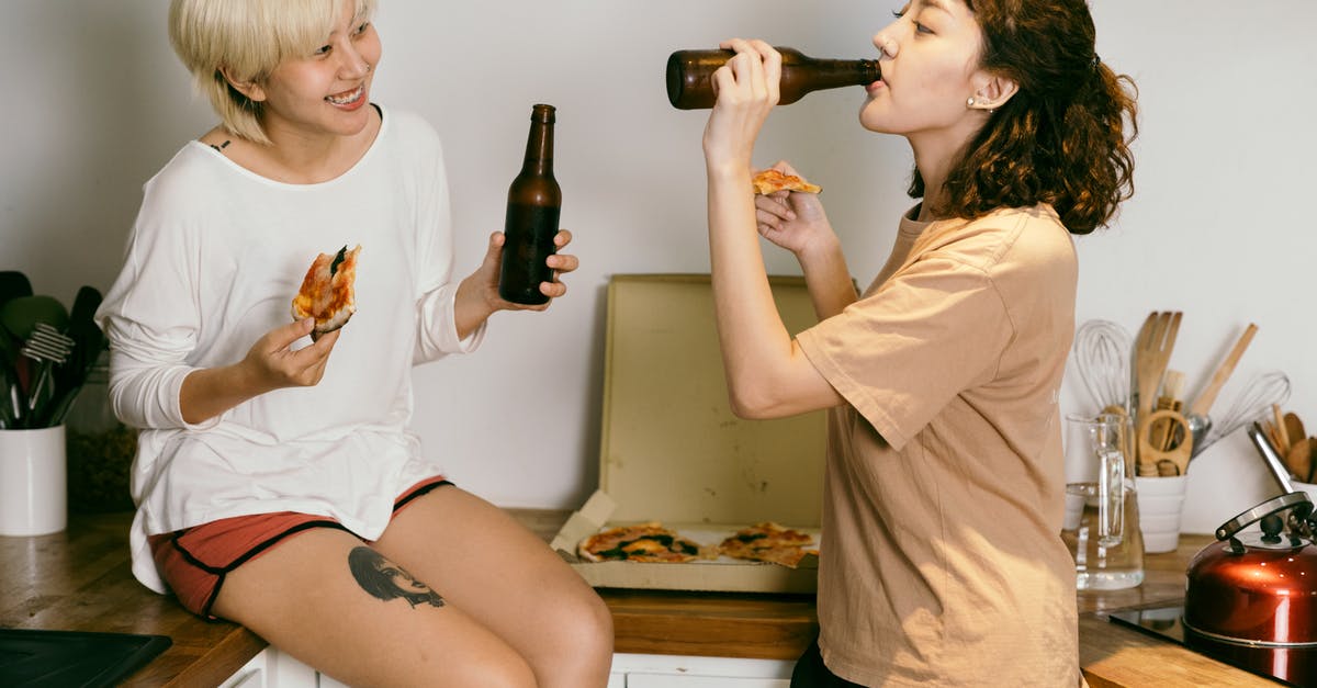 What are other uses for a pizza stone? - Happy tattooed blond lady eating pizza while looking at female friend in casual wear drinking beverage from bottle near kitchen utensils in apartment