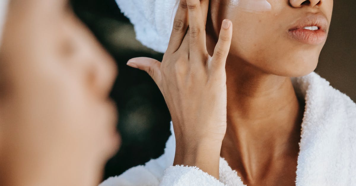 What does make ice cream smooth and silky? - Crop unrecognizable young ethnic female in terry robe applying moisturizing cream on cheek while reflecting in mirror