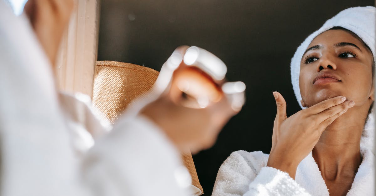 What does make ice cream smooth and silky? - African American young female in white bathrobe applying moisturizing cream on face while standing in bathroom