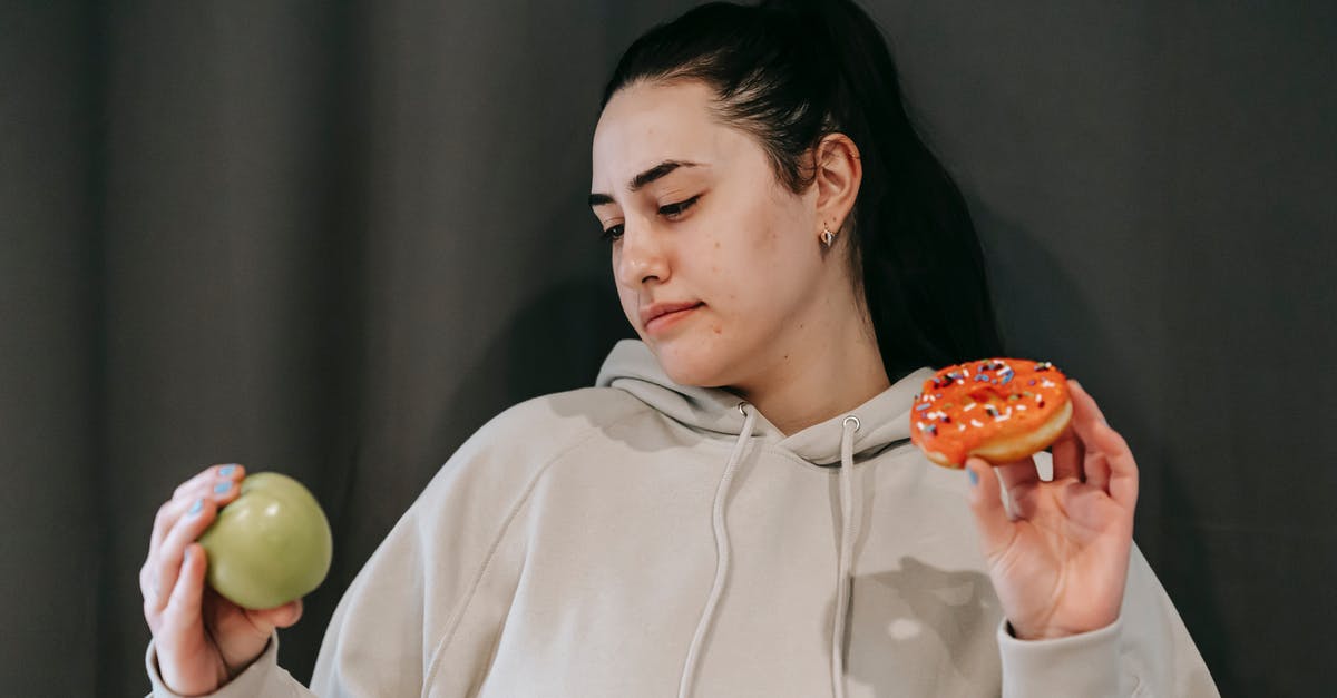 What is the difference between doughnut and krapfen? - Pensive female wearing casual hoodie deciding between healthy green apple and sweet doughnut in studio