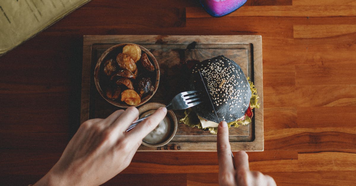 What is the point of mustard-frying burger patties? - Top view of crop anonymous person cutting appetizing black burger served on wooden board with sauce and fried potatoes