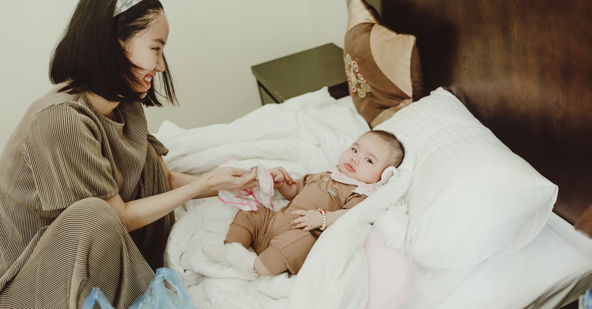 What is the reason to coddle an egg? - Young smiling Asian female mom in home clothes coddling with baby in bodysuit lying on white coverlet on bed