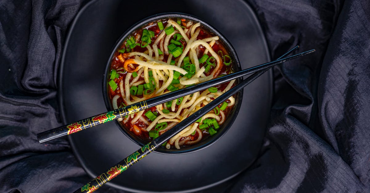 What is this Asian dish made with boiled vegetables?  - Top view of black bowl with Asian soup with noodle and green onion served with bamboo chopsticks on black tablecloth