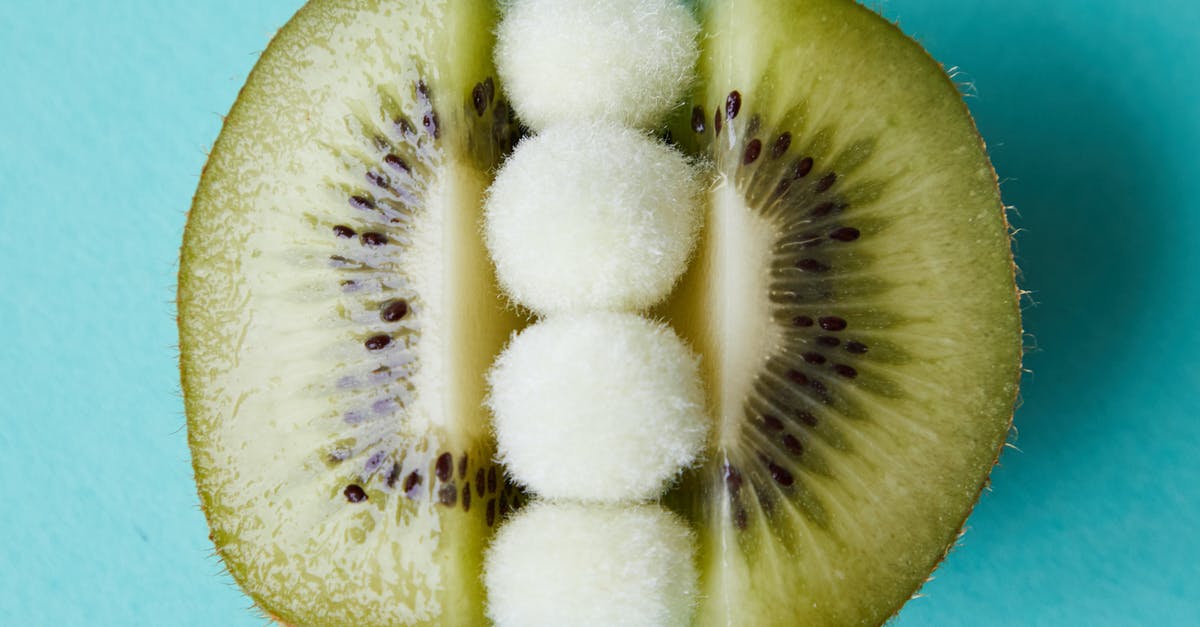 What is this small green fruit called? - Fur pom poms between halves of kiwi on blue surface