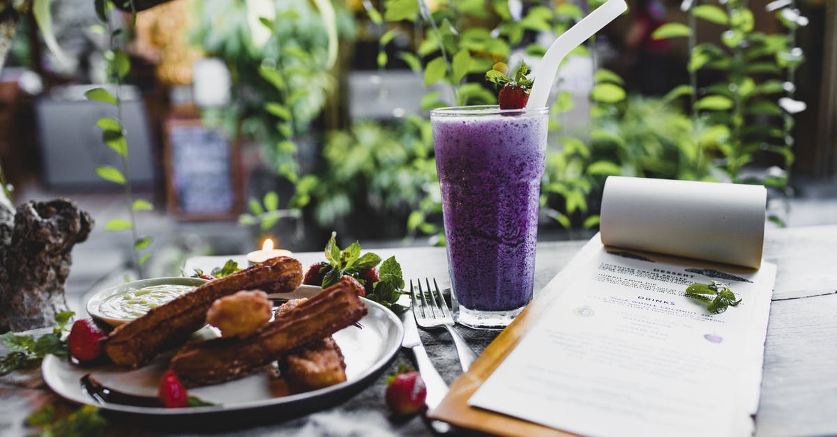 What is this Spanish dessert? - Glass of yummy blueberry milkshake with straw near plate of churros with fresh strawberries and menu with cutlery on table in front of climbing plant in restaurant