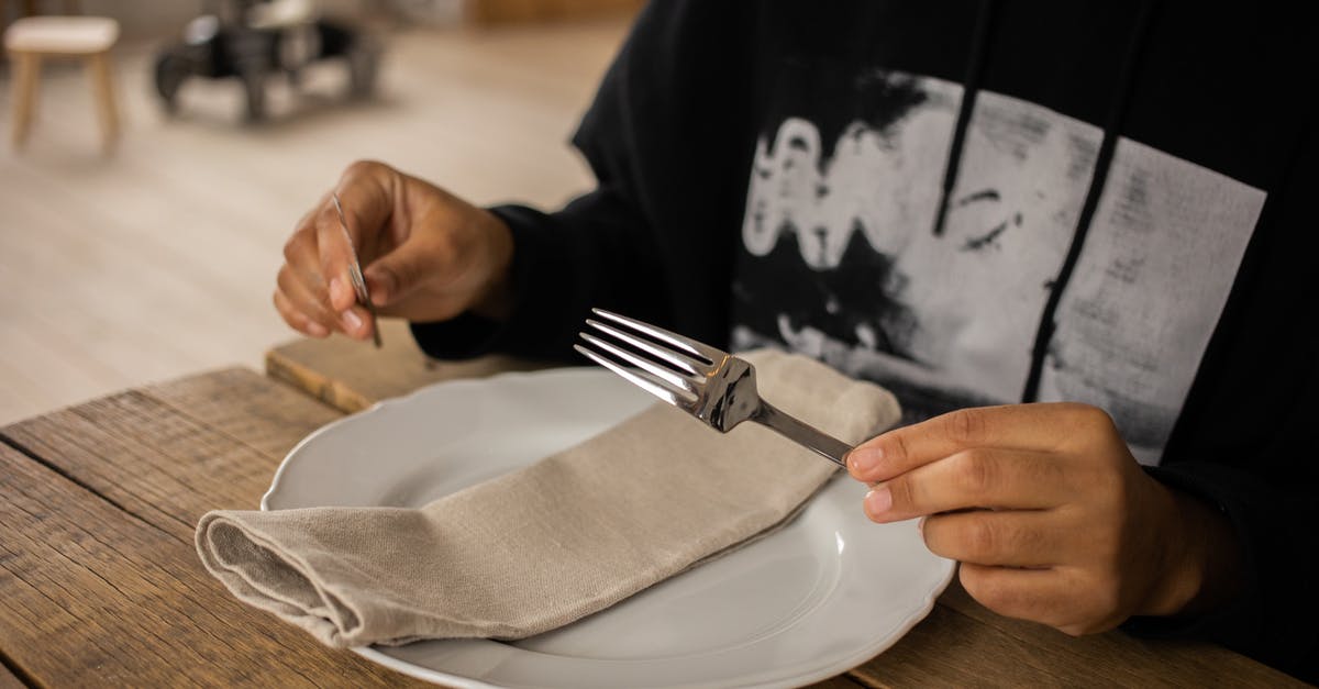 What precautions should be taken while cleaning silver utensils? - Crop anonymous person sitting at wooden table with fork and knife in hands near white plate with napkin on blurred background