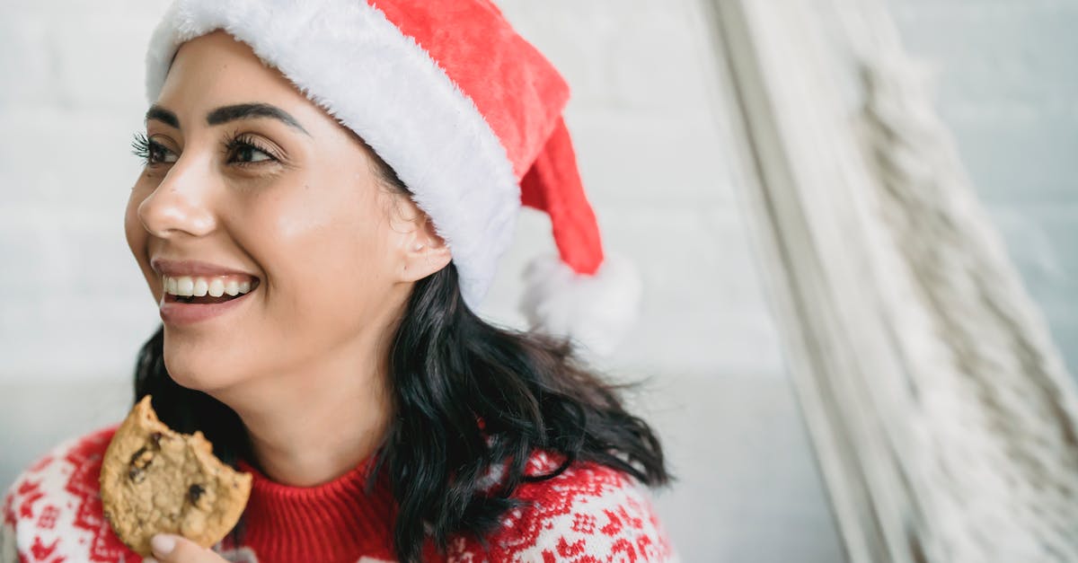 What should I look for in a cookie jar? - Smiling young woman in Santa hat and sweater eating chocolate chip cookie and looking away in light room