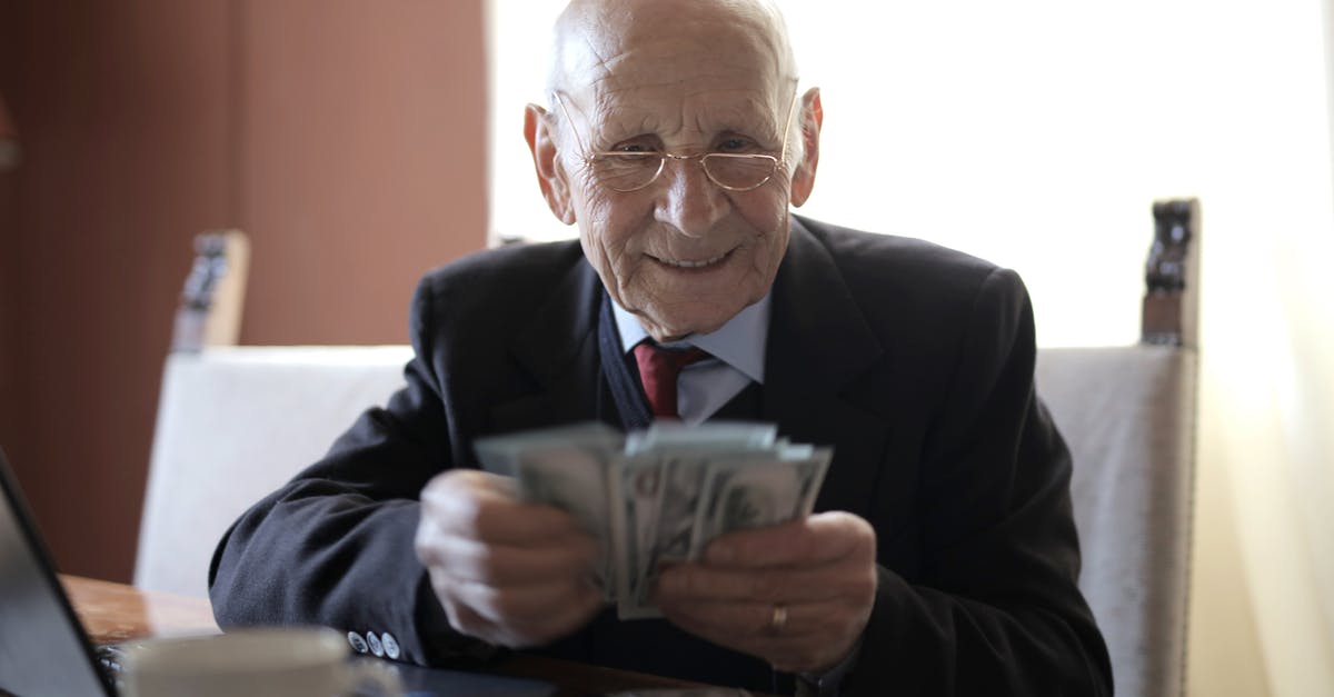 What to do with old coffee beans? - Positive senior businessman in formal suit and eyeglasses counting money bills while sitting at wooden table with cup of beverage and near opened laptop