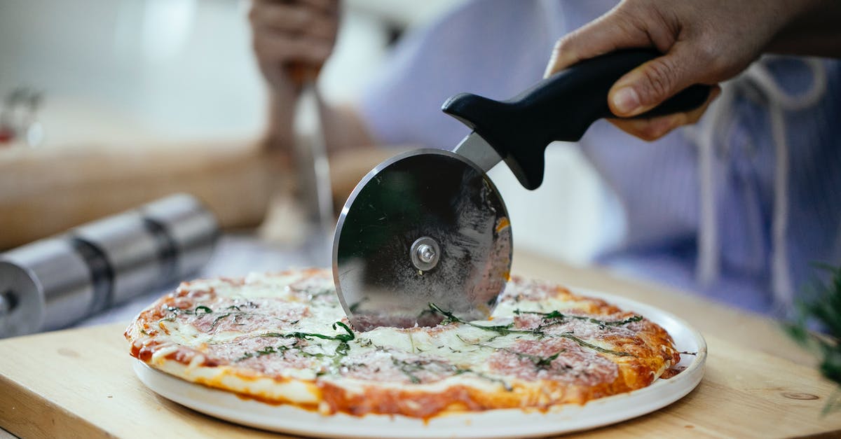 When to add fresh basil to a tomato sauce? - Unrecognizable person cutting pizza with melted cheese and herbs using pizza cutter in kitchen