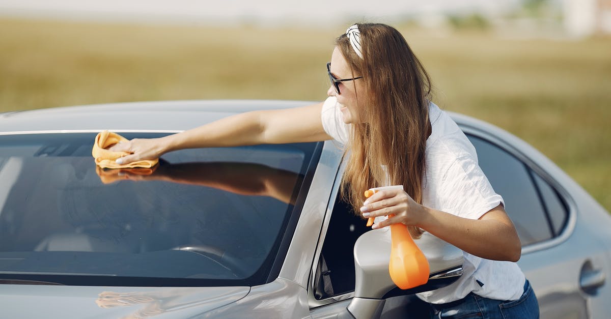 When to spray or when to use olive oil? - Side view of cheerful female driver in sunglasses and casual clothes cleaning windshield of modern car with microfiber cloth and spray bottle against green field