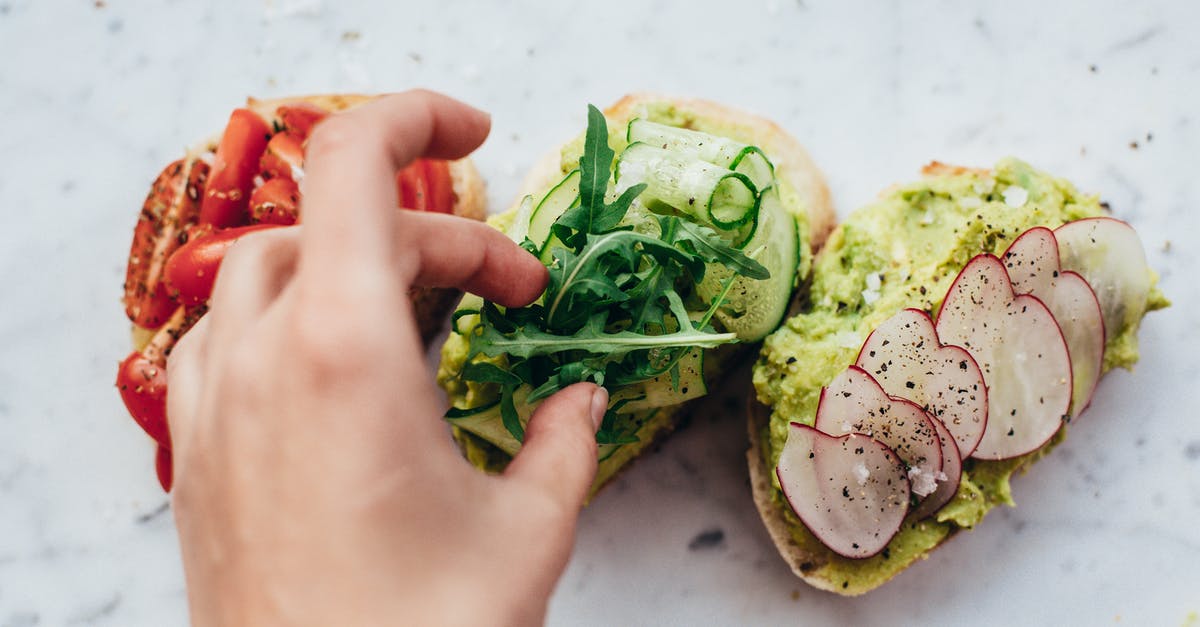 Which ingredient in Italian bread gives it that great taste? - From above of crop anonymous person adding arugula on bruschetta with cucumber and avocado placed near slices of bread with tomatoes and radish