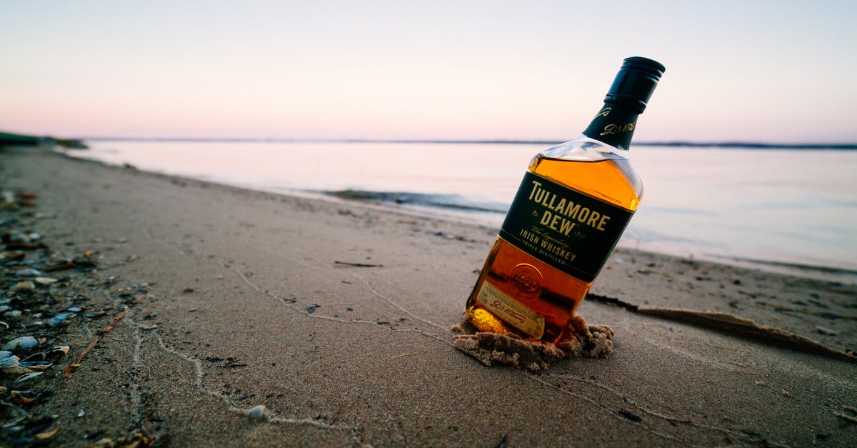 Whiskey and Water - Ground level of strong elite alcoholic drink in transparent glass bottle on wet sandy shore near calm endless ocean under sky at sunset