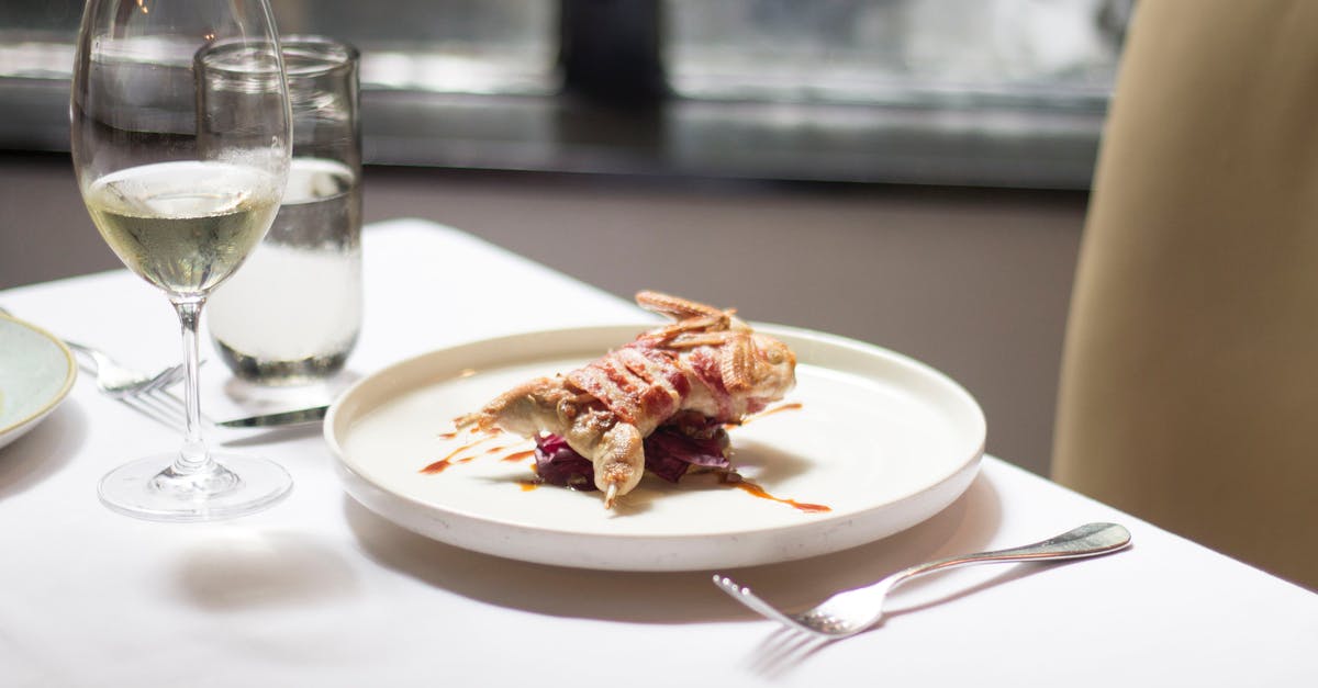 White goo from chicken ruins the dish - Tablecloth on table with delicious plate with small roasted bird covered in bacon with sauce near glasses with wine and water near forks and knife in light restaurant near chair