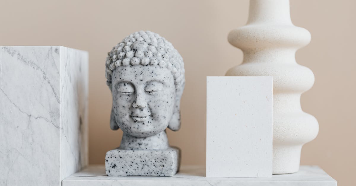 white spots on top of indian pickle - Granite bust of Buddha placed near white ceramic vase of creative geometric shape and blank card on white marble shelf against beige wall
