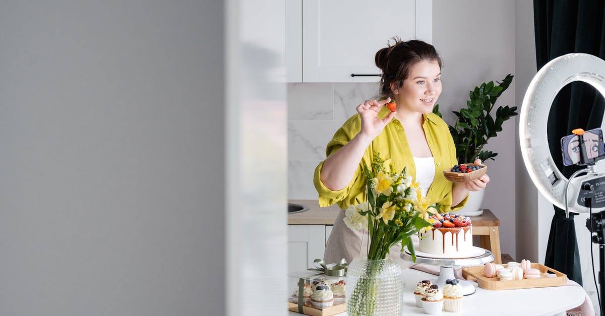 Why is my cupcake brownie like? - Delighted young female influencer in casual clothes smiling and demonstrating fresh berries while decorating appetizing cake during recording vlog on smartphone in kitchen