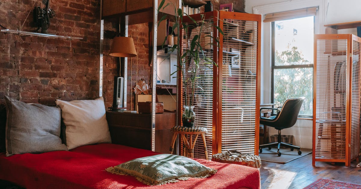 Why not cover the pot? - Comfortable bed with red cover and pillows placed near brick wall in cozy room with isolated workspace decorated with houseplants near window