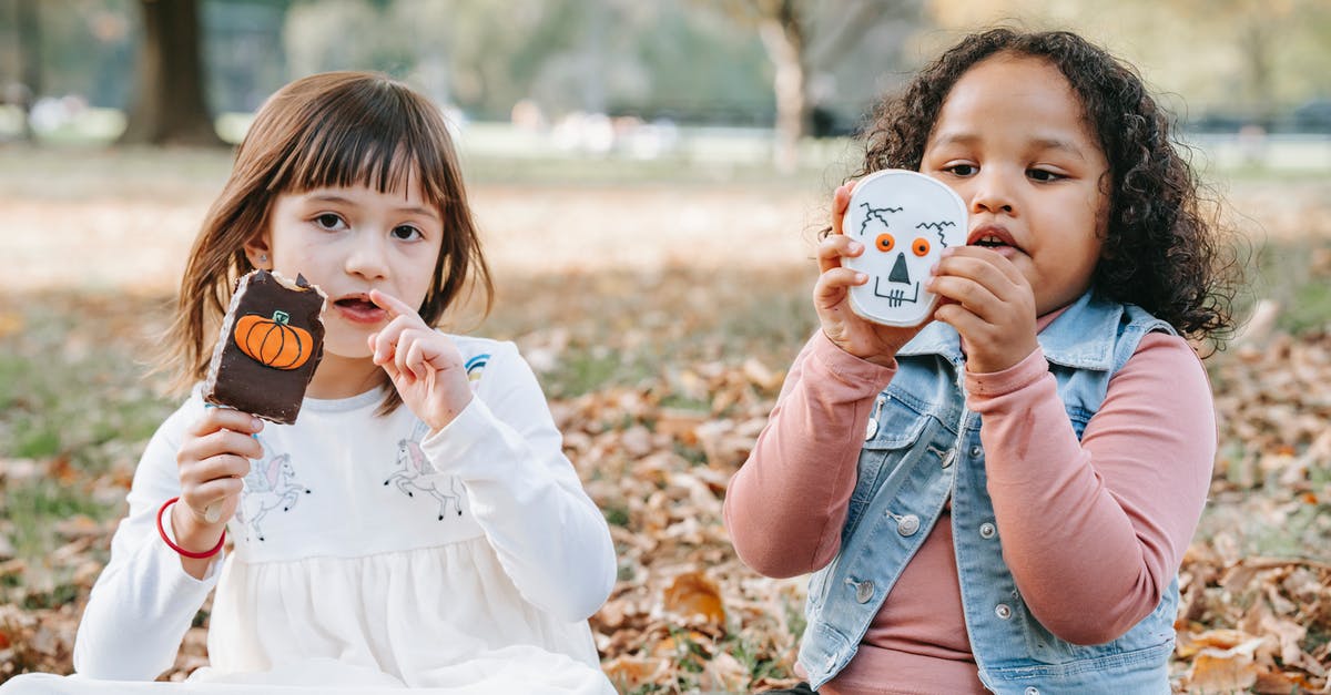 Why should I rest gingerbread dough? - Adorable little multiethnic girls in casual outfits sitting on ground in park with sweets decorated for Halloween